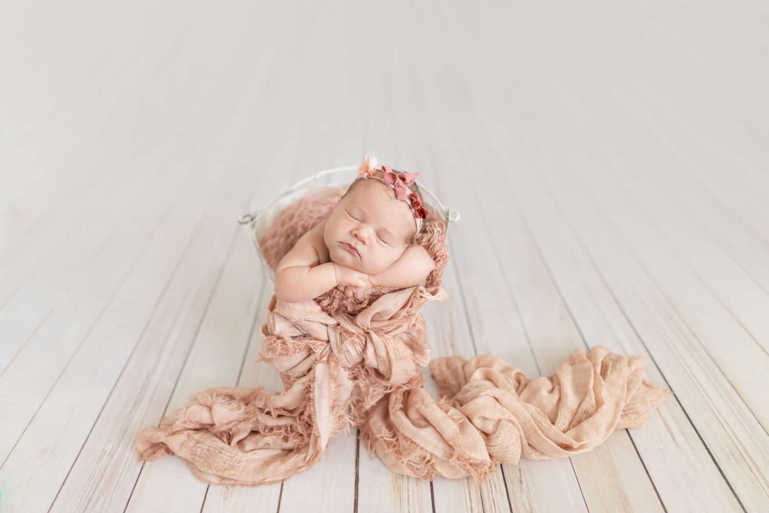 Newborn baby sleeping peacefully in a pink wrap and floral headband, posed in a bowl on white wood floors.
