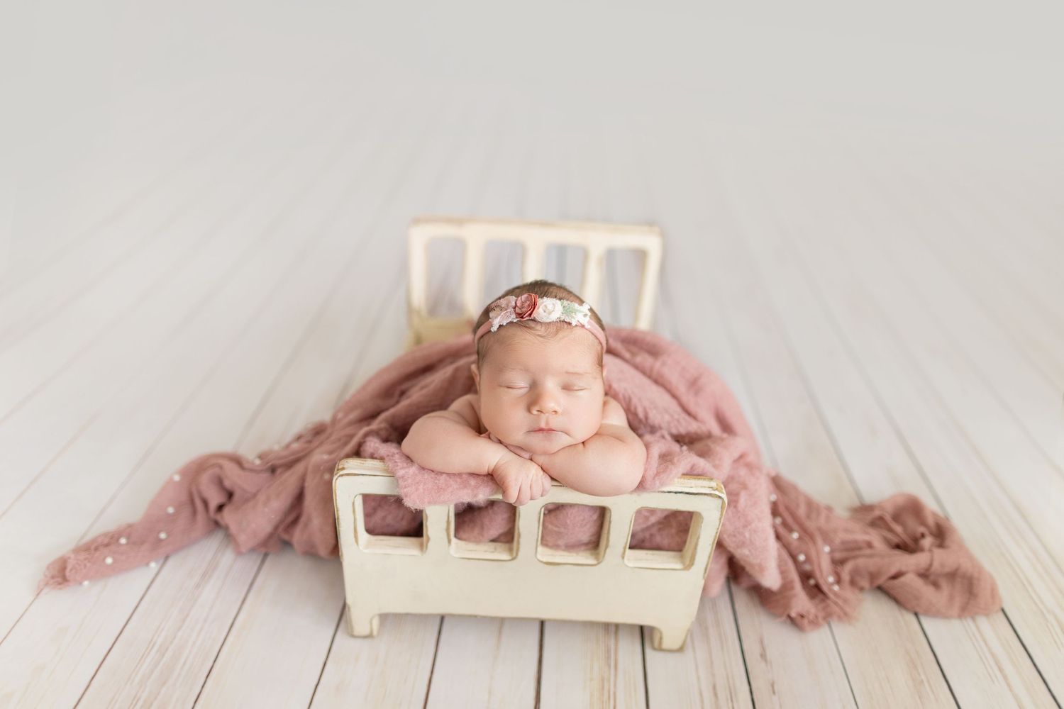 Newborn baby girl sleeping peacefully in a tiny white bed with pink blanket and floral headband.