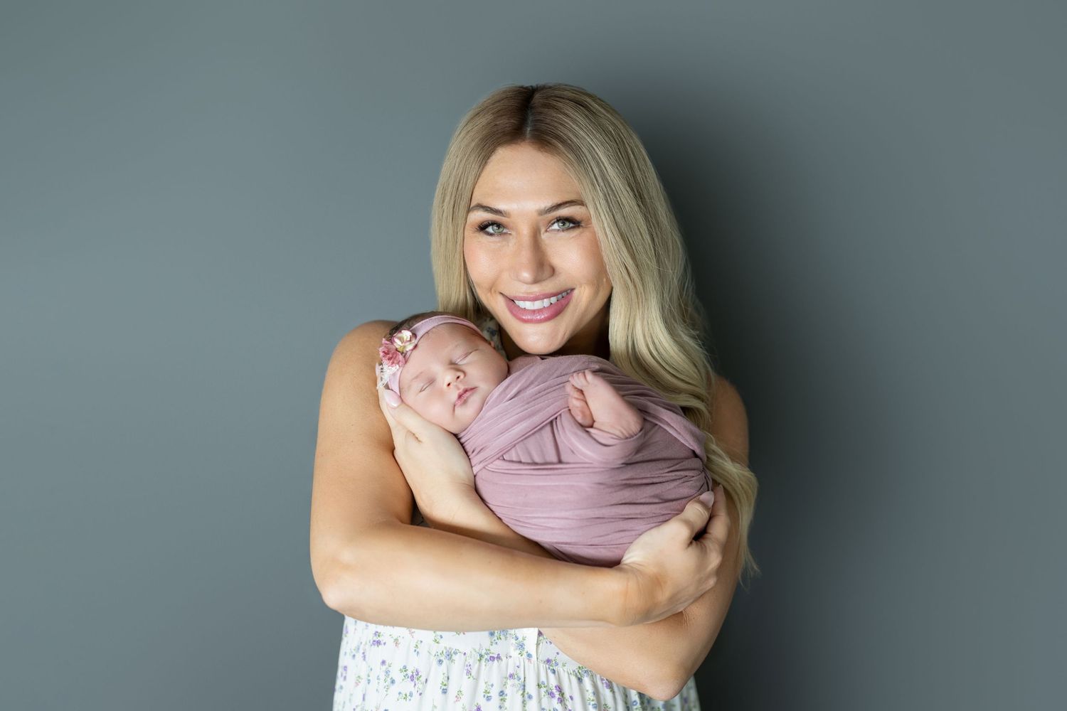 Smiling blonde woman holding a swaddled newborn baby in a pink wrap against a gray background.