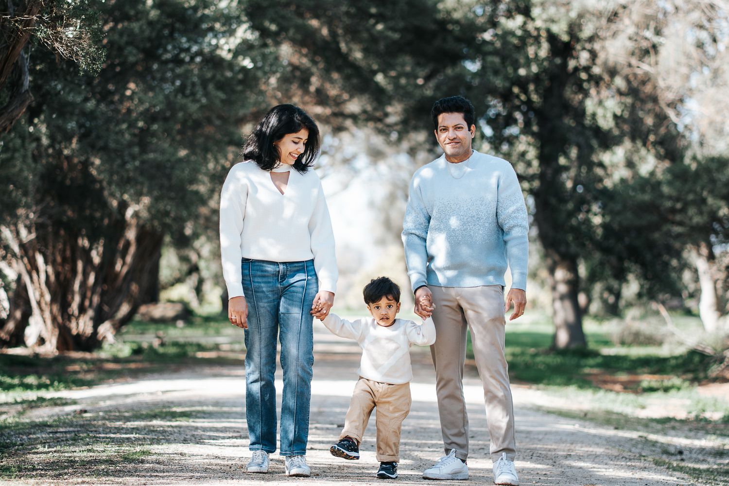 Family of three walking hand-in-hand on a tree-lined path in a sunny park in Melbourne taken by a Melbourne family photographer.