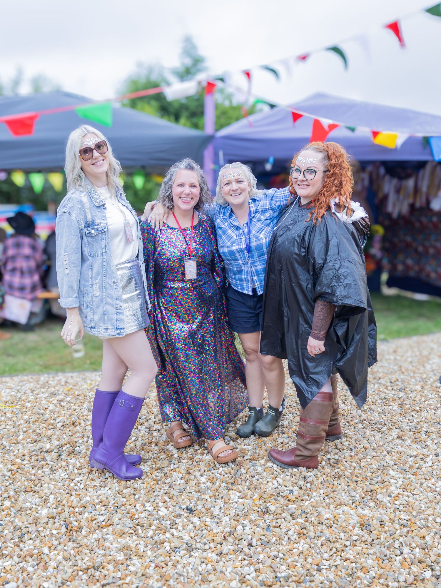 Four women posing together at an outdoor festival with colorful bunting flags and market stalls in the background.