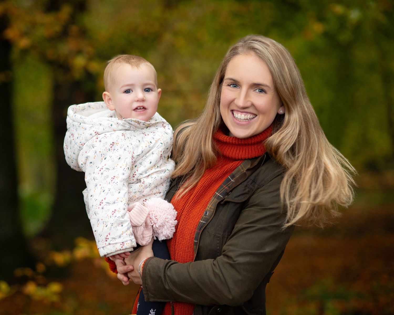 Smiling mother in a red scarf holds a bundled baby outdoors in an autumn park; both face the camera, creating a warm, joyful portrait.