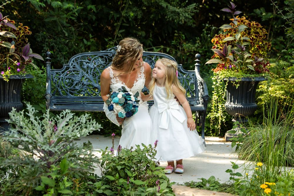 Bride in white gown holding teal bouquet shares a sweet moment with flower girl in garden.