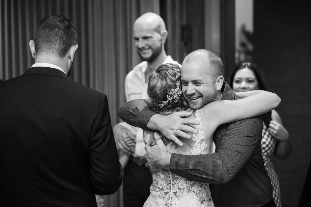 Black and white wedding photo of bride and groom sharing a warm embrace with guests smiling in background.