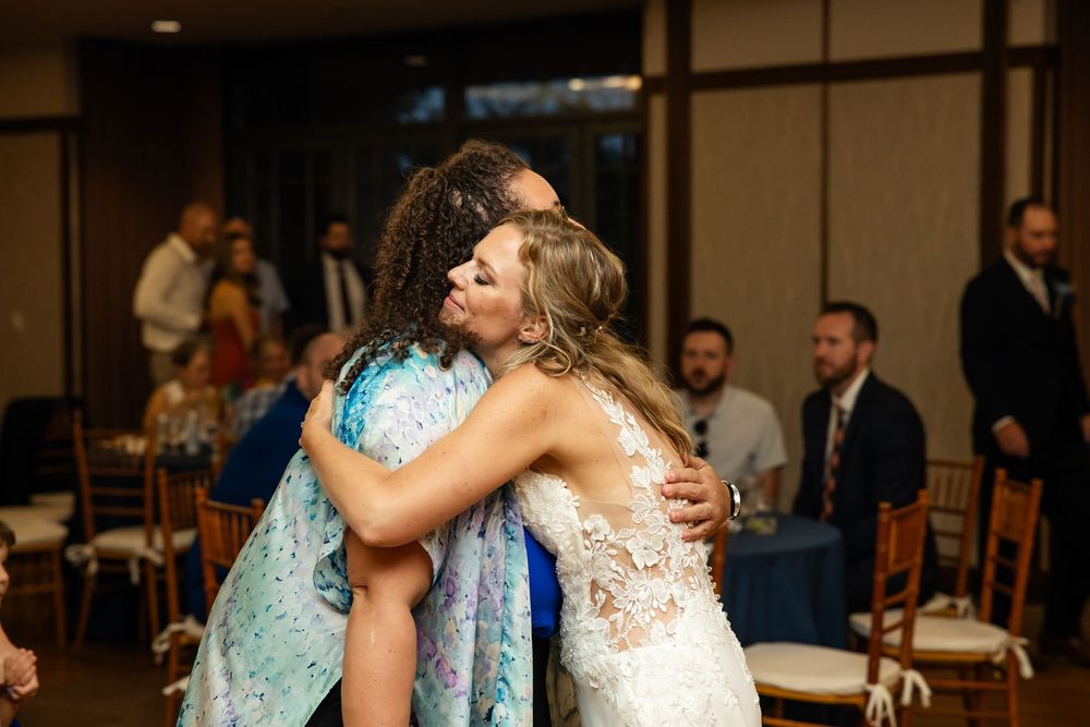 Bride in lace gown shares warm embrace with guest in floral dress at elegant wedding reception.