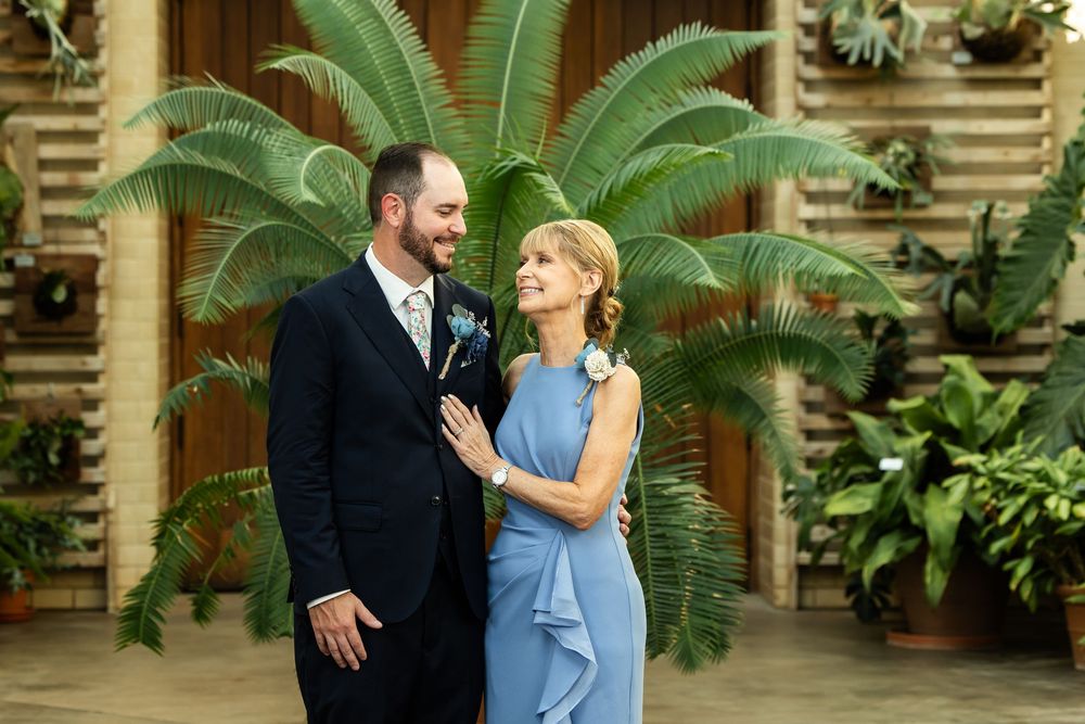 Couple in elegant attire poses before lush tropical greenery, man in dark suit, woman in blue ruffled dress.