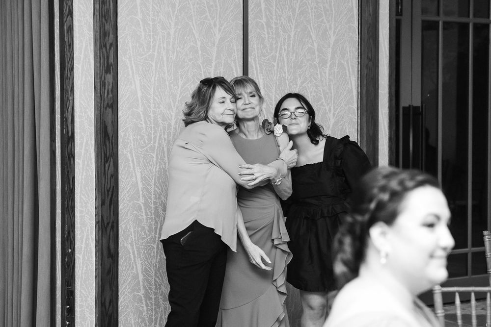 Three women share a warm group hug at a formal event in this elegant black and white photo.