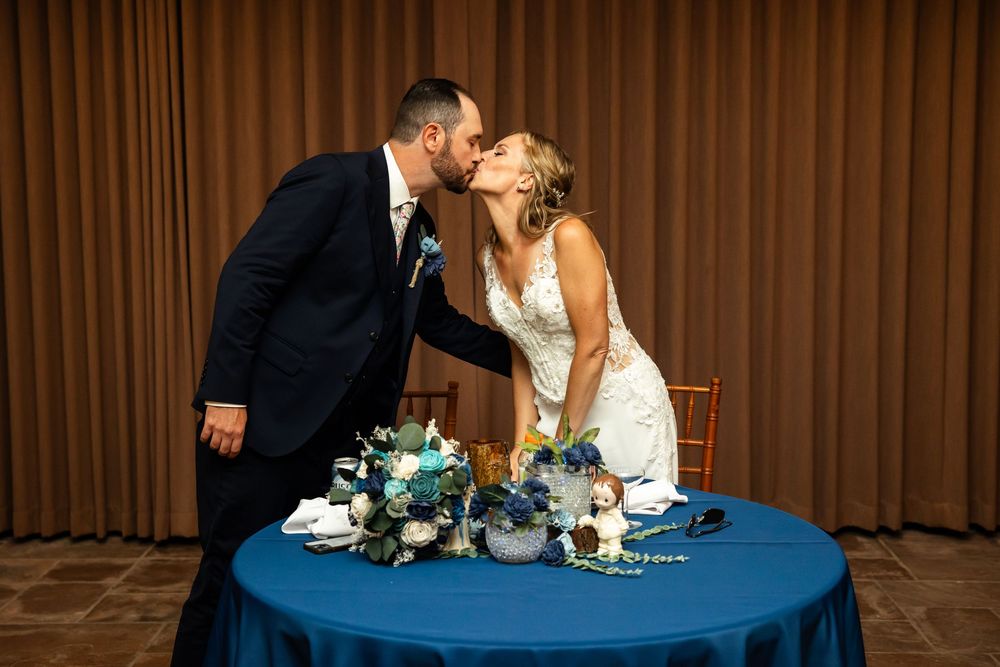 Bride and groom share a kiss at their sweetheart table adorned with blue linen and floral centerpiece.
