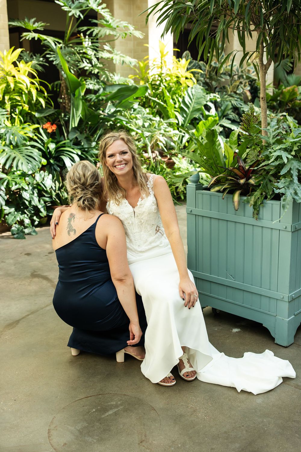 Bride in white gown smiles as bridesmaid in black dress adjusts her shoes amid lush tropical greenery.