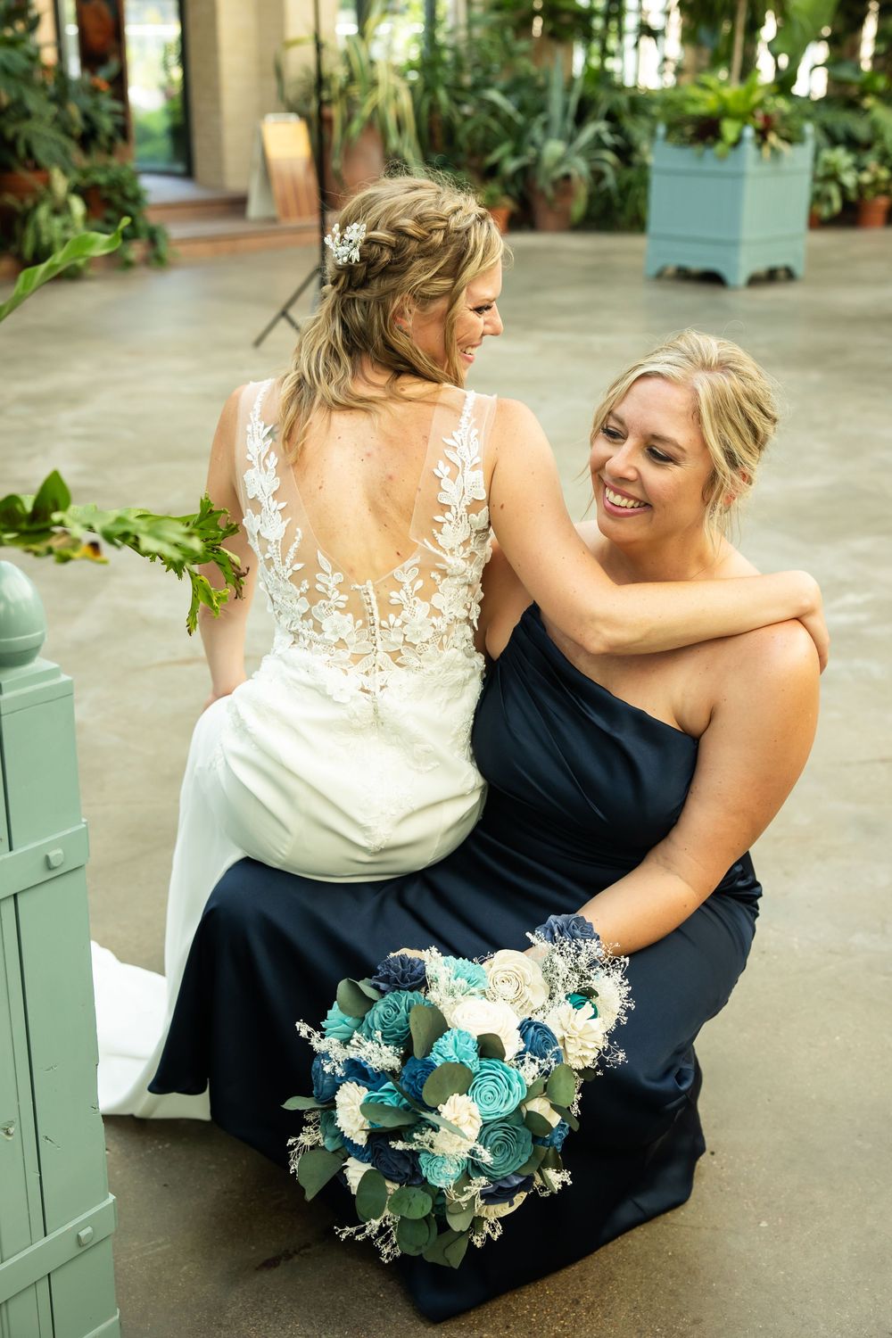 Bride in lace backless gown with bridesmaid in navy dress holding teal bouquet, smiling together at wedding venue.