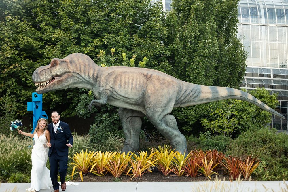 Wedding couple poses in front of a large T-Rex dinosaur statue surrounded by colorful plants outdoors.