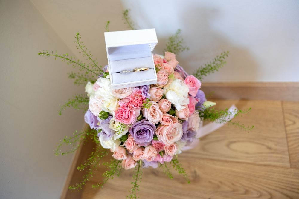 A bridal bouquet of pink, white, and purple roses with two gold wedding rings in a white box on top.