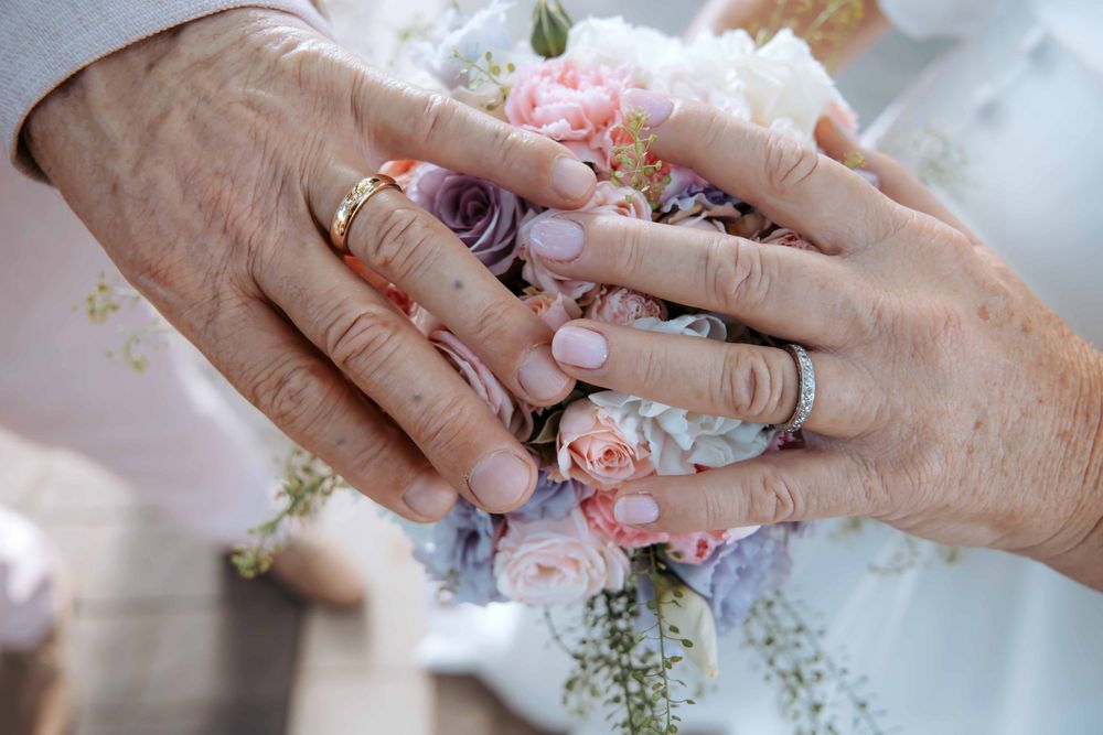 Elderly couple's hands with wedding rings resting on a pastel bridal bouquet of pink and purple flowers.