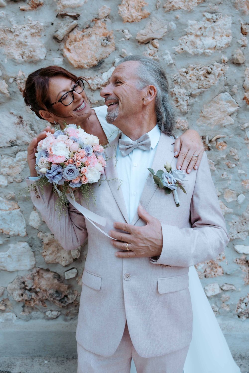Elegant older couple in wedding attire smiling, groom in pink suit with boutonniere, bride holding pastel bouquet against stone wall.
