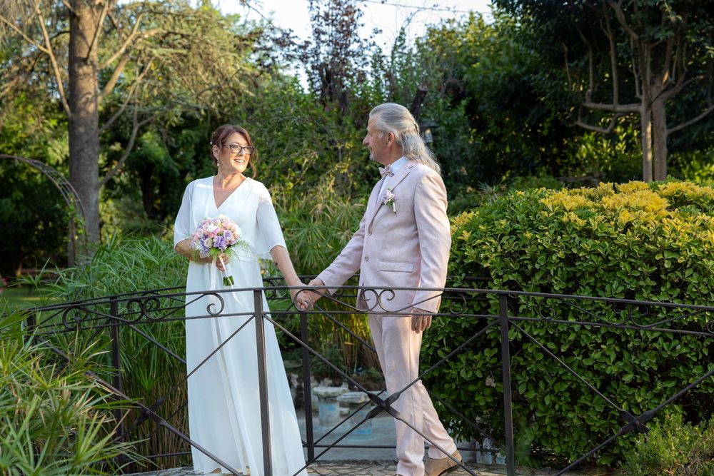Bride and groom holding hands on a garden bridge, bride in white gown holding bouquet, groom in light pink suit.