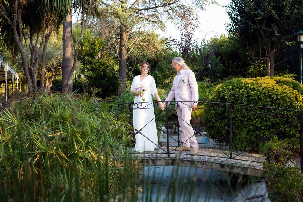 Bride and groom holding hands on a wooden bridge surrounded by lush green gardens on their wedding day.