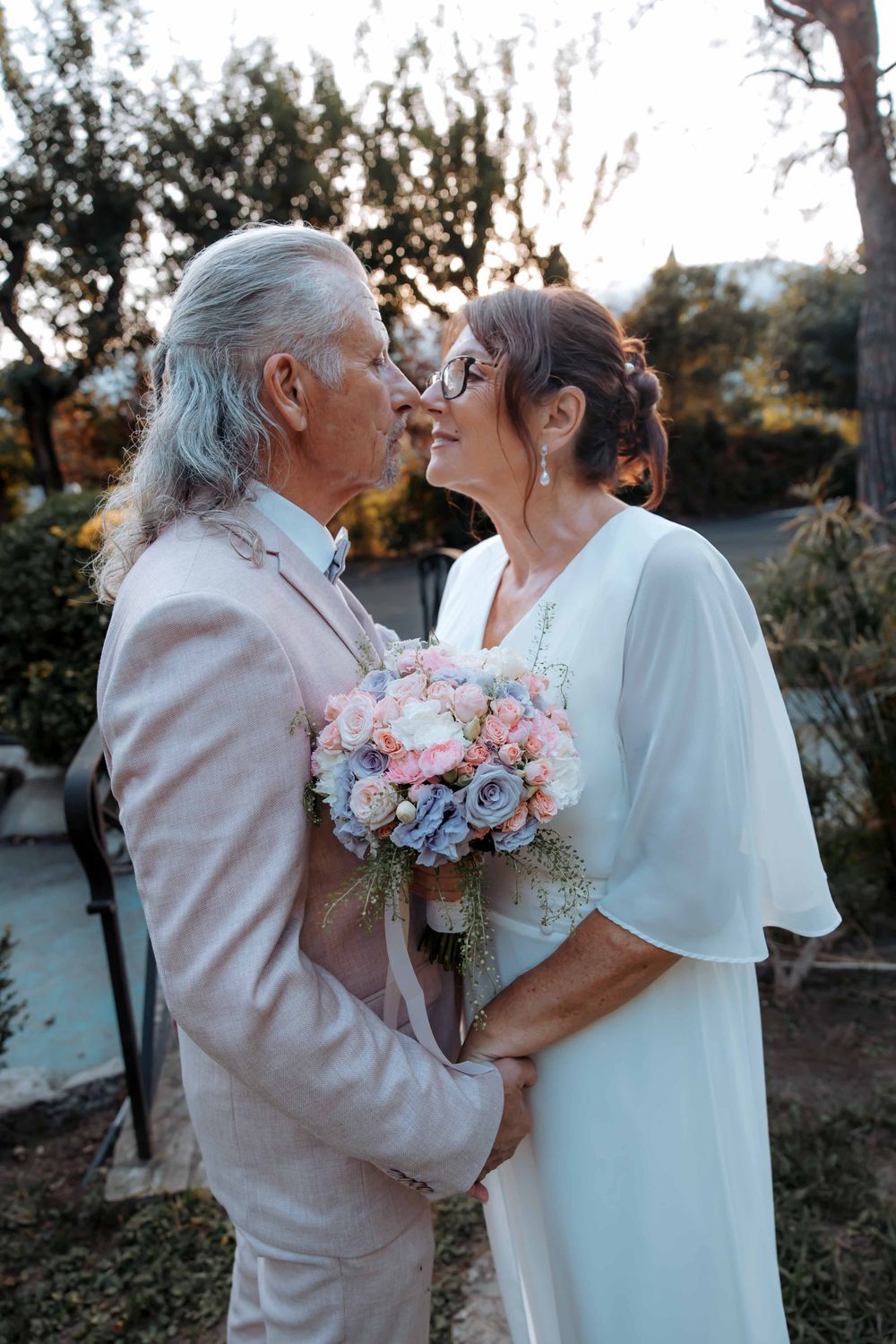 Elderly couple in wedding attire sharing a romantic moment, bride holding pastel bouquet outdoors.