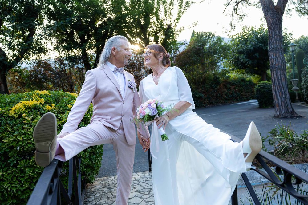Joyful wedding couple in a garden, bride in white gown holding bouquet, groom in pink suit, both kicking legs up playfully.