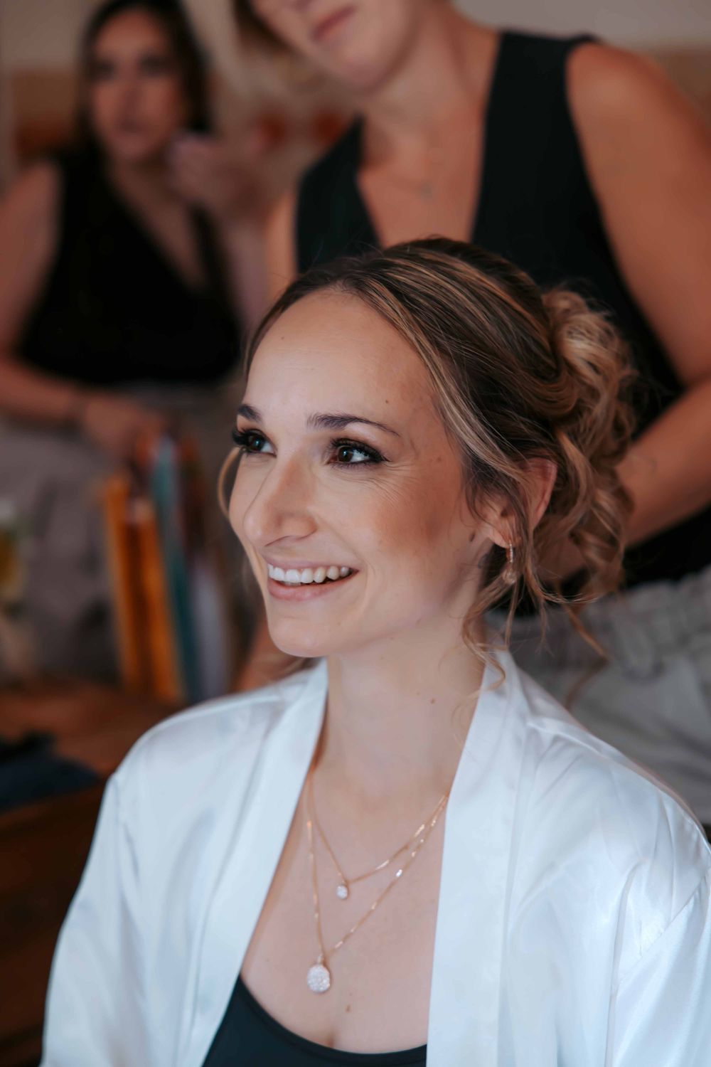 Smiling bride in white robe with updo hairstyle and layered necklace getting ready with bridesmaids in background.