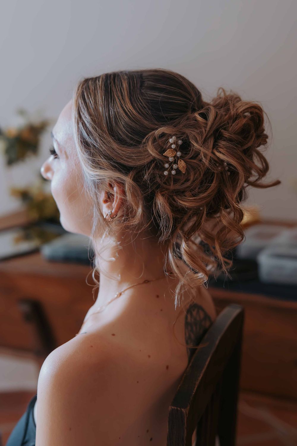 Woman with elegant curly updo hairstyle adorned with floral hair pins, seated in chair showing back view.