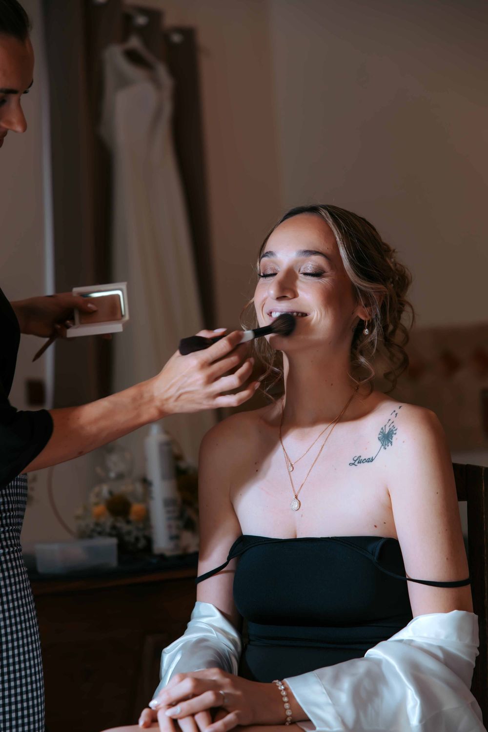 A makeup artist applying powder to a seated woman in a black strapless dress with a floral tattoo on her shoulder.