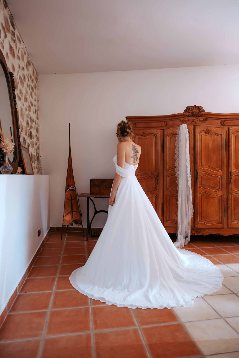 Bride in elegant white backless wedding gown with long train standing in rustic room with wooden armoire.