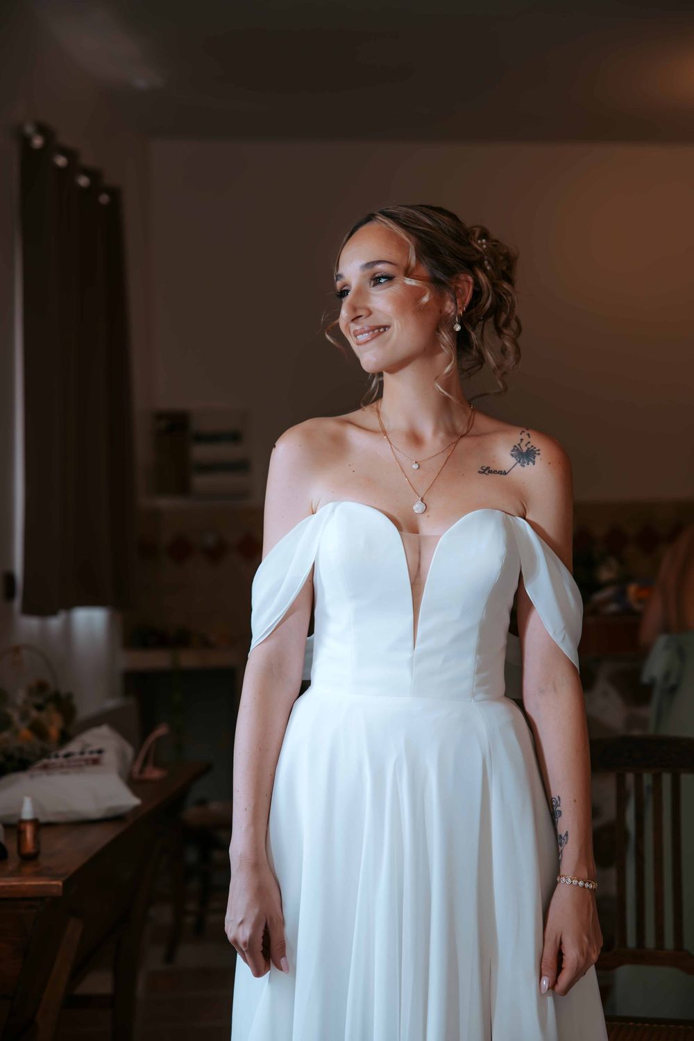 Smiling bride in off-shoulder white gown with plunging neckline, updo hairstyle, and shoulder tattoo in warm indoor setting.