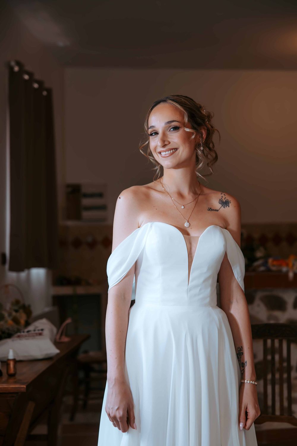 Smiling bride in off-shoulder white wedding gown with deep V-neckline and shoulder tattoo posing indoors.