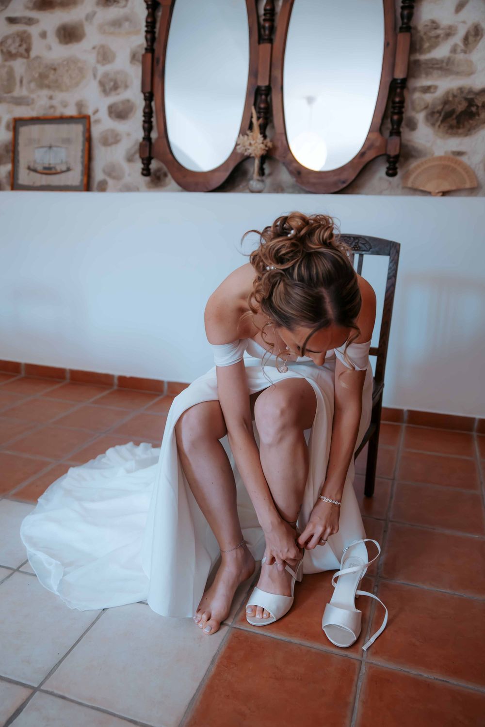 Bride in white off-shoulder wedding dress sitting on chair, fastening strappy heels, with ornate mirrors on stone wall.