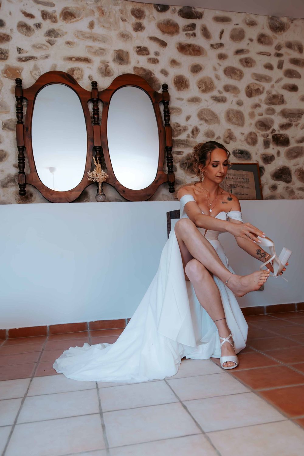Bride in white wedding gown adjusting her heels near a rustic stone wall with ornate oval mirrors.