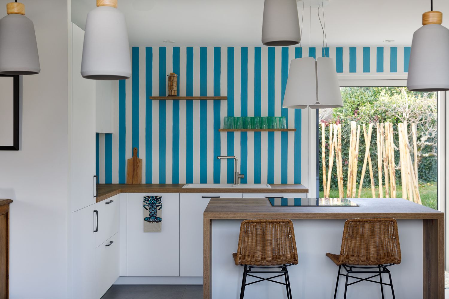 Cuisine lumineuse agrémentée d'un papier peint audacieux à rayures bleues et blanches, de meubles blancs, d'un îlot en bois et de tabourets