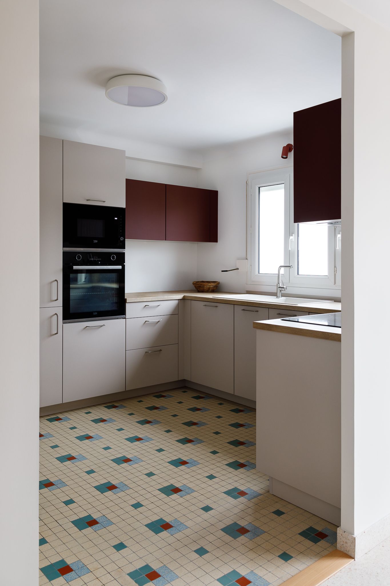 Cuisine moderne en L dotée de meubles hauts bordeaux, de meubles bas beiges, d'un double four et d'un sol en carrelage à motifs géométriques