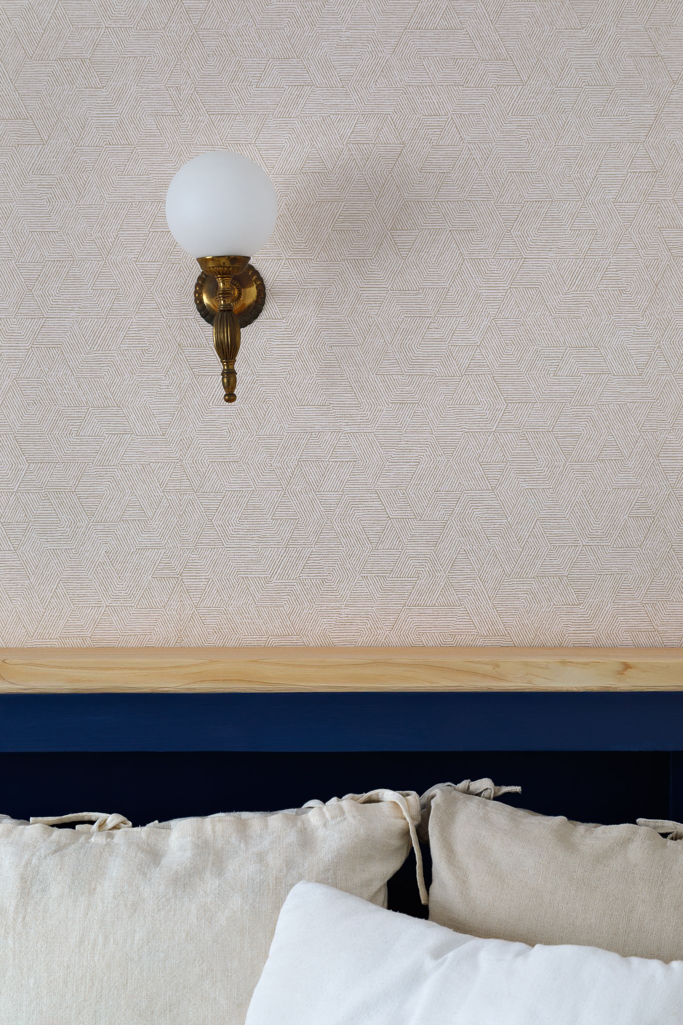 Détail de la chambre : applique murale en laiton avec abat-jour globe au-dessus d’une tête de lit bleu marine avec coussins en lin.