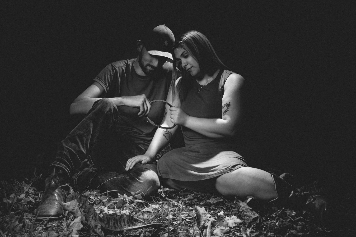 Black and white photo of a man and woman sitting together on the ground in a dark, moody outdoor setting.