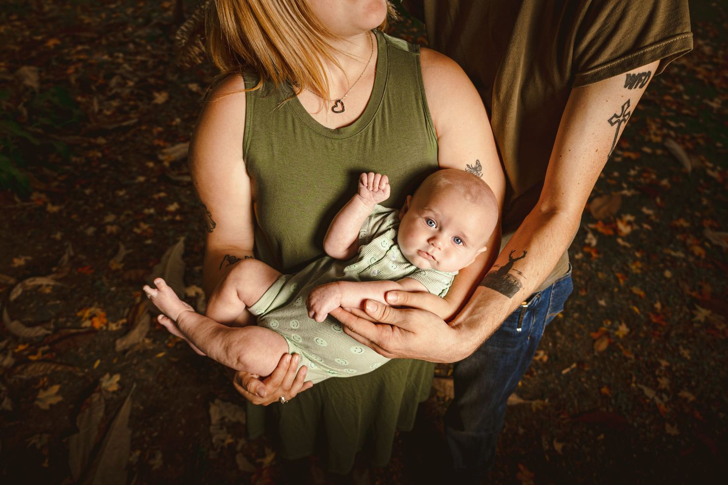 Parents cradling their newborn baby dressed in green onesie outdoors among autumn leaves.