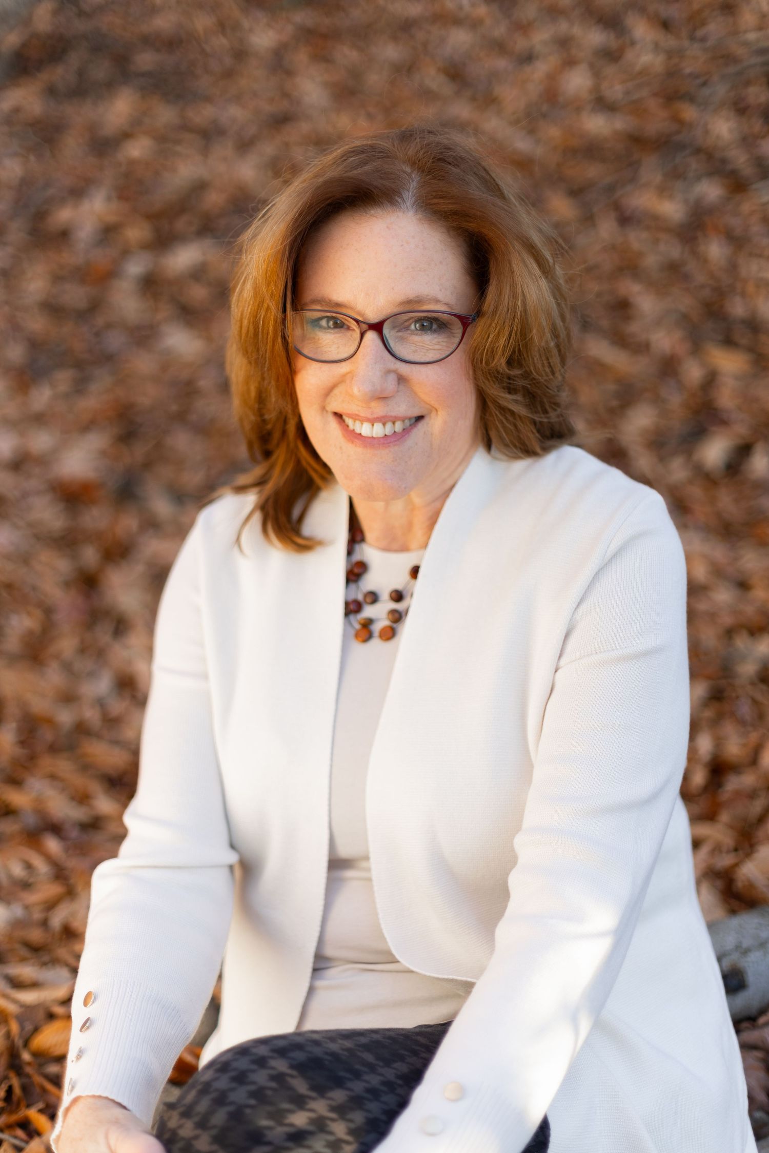 Smiling middle-aged woman with red hair and glasses wearing a white blazer, seated outdoors among autumn leaves.