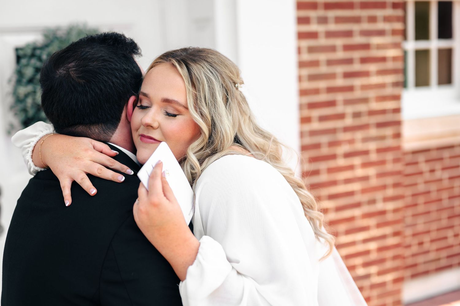 A bride in a white dress embraces her groom in a black suit, sharing an emotional moment near a brick building.
