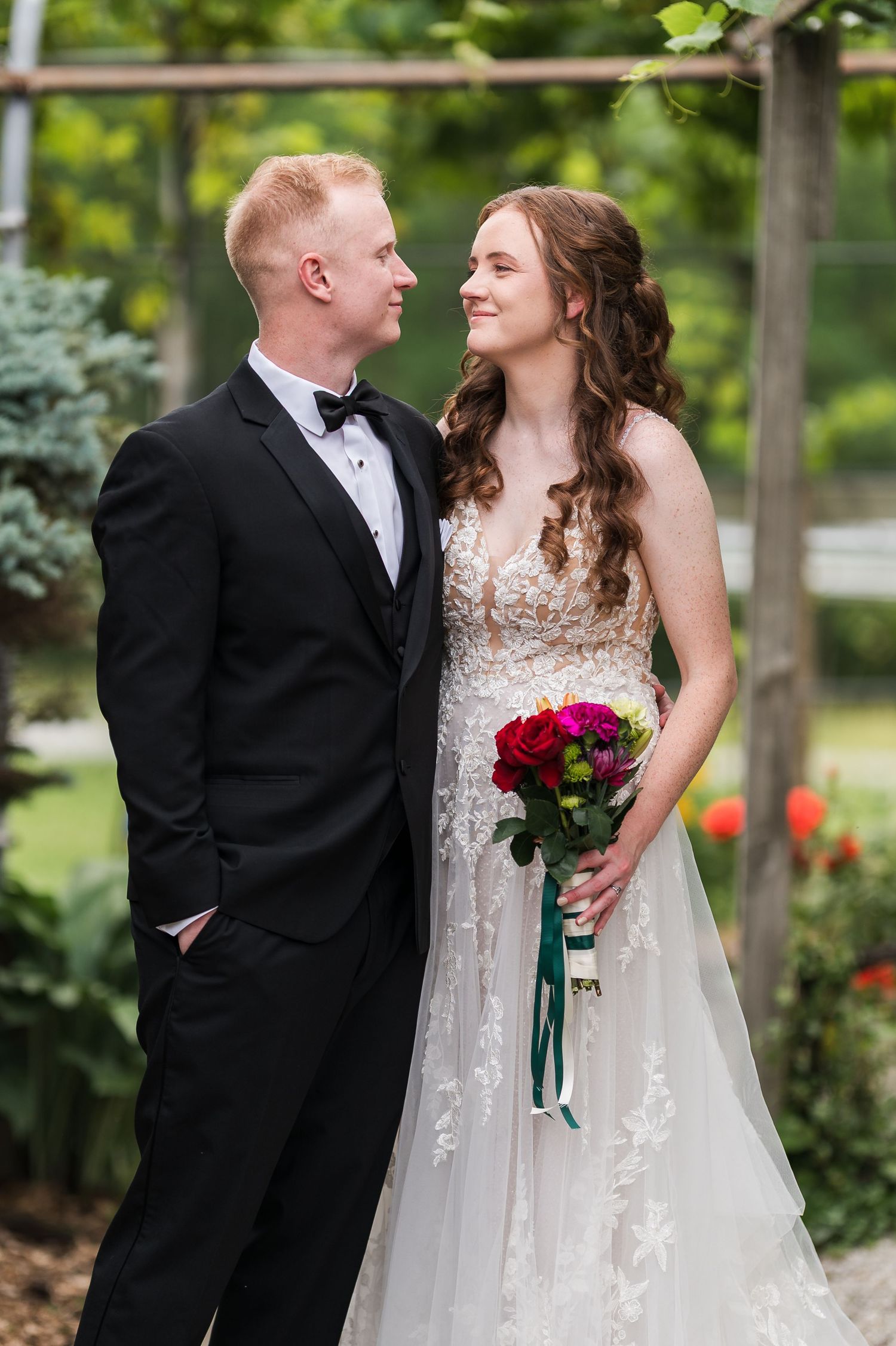 A bride and groom share a romantic moment outdoors, she holds a vibrant bouquet in a lace gown as he wears a black tuxedo.