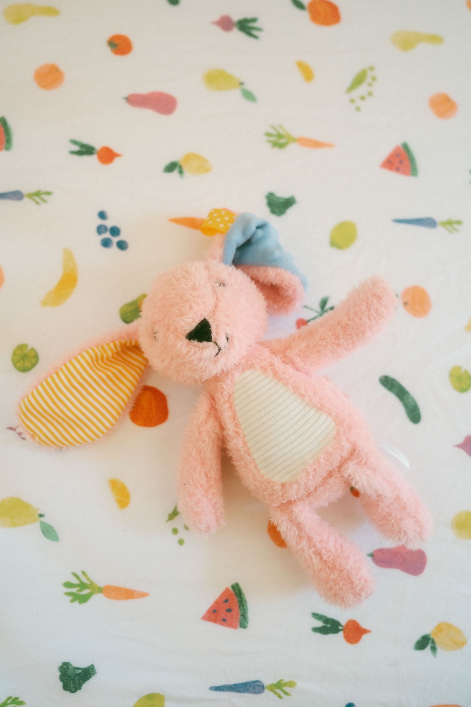 Pink bunny stuffed animal toy laying on a white fabric with colorful fruit and vegetable print pattern.