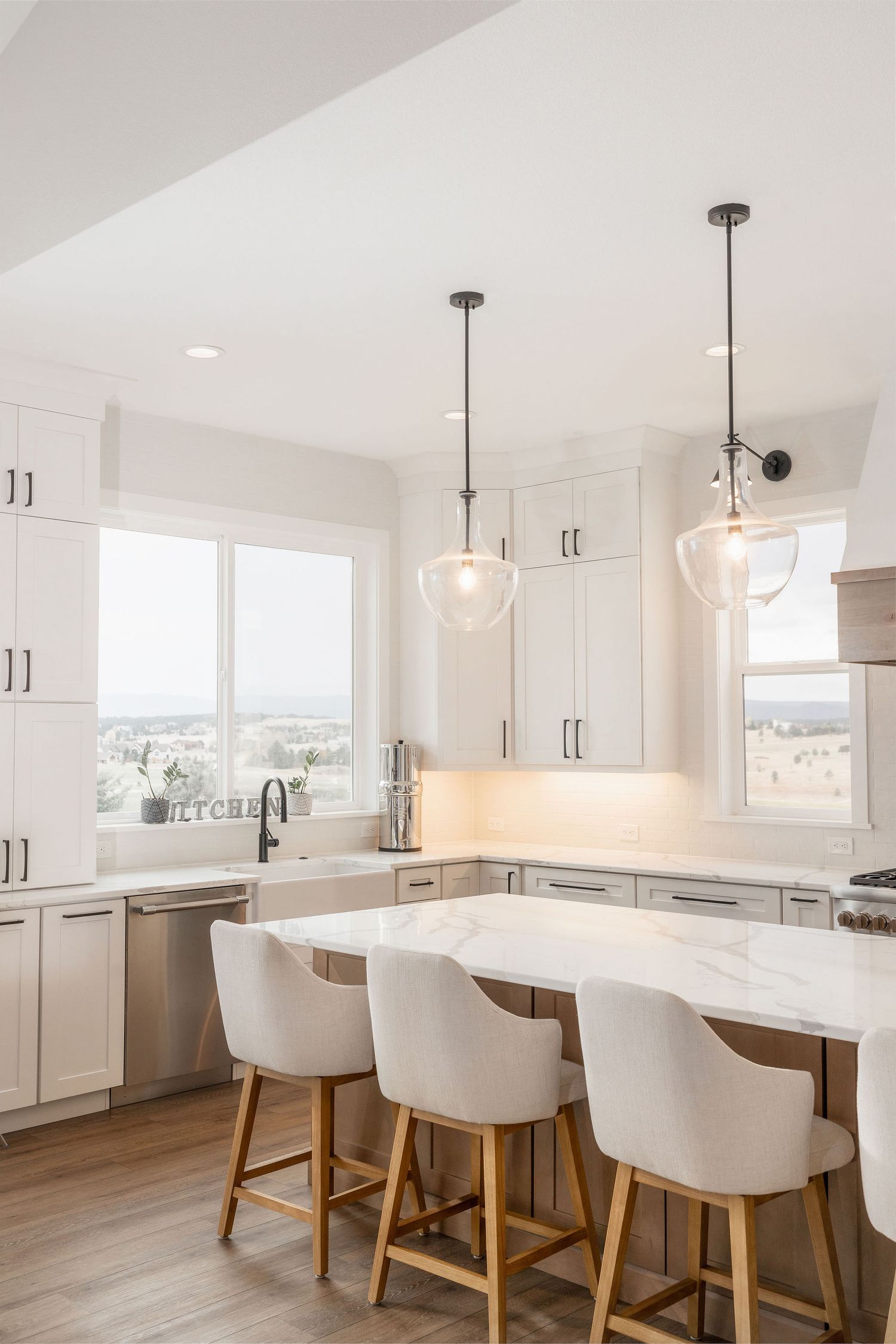 Modern white kitchen with marble island, bar stools, pendant lights, and scenic window views.
