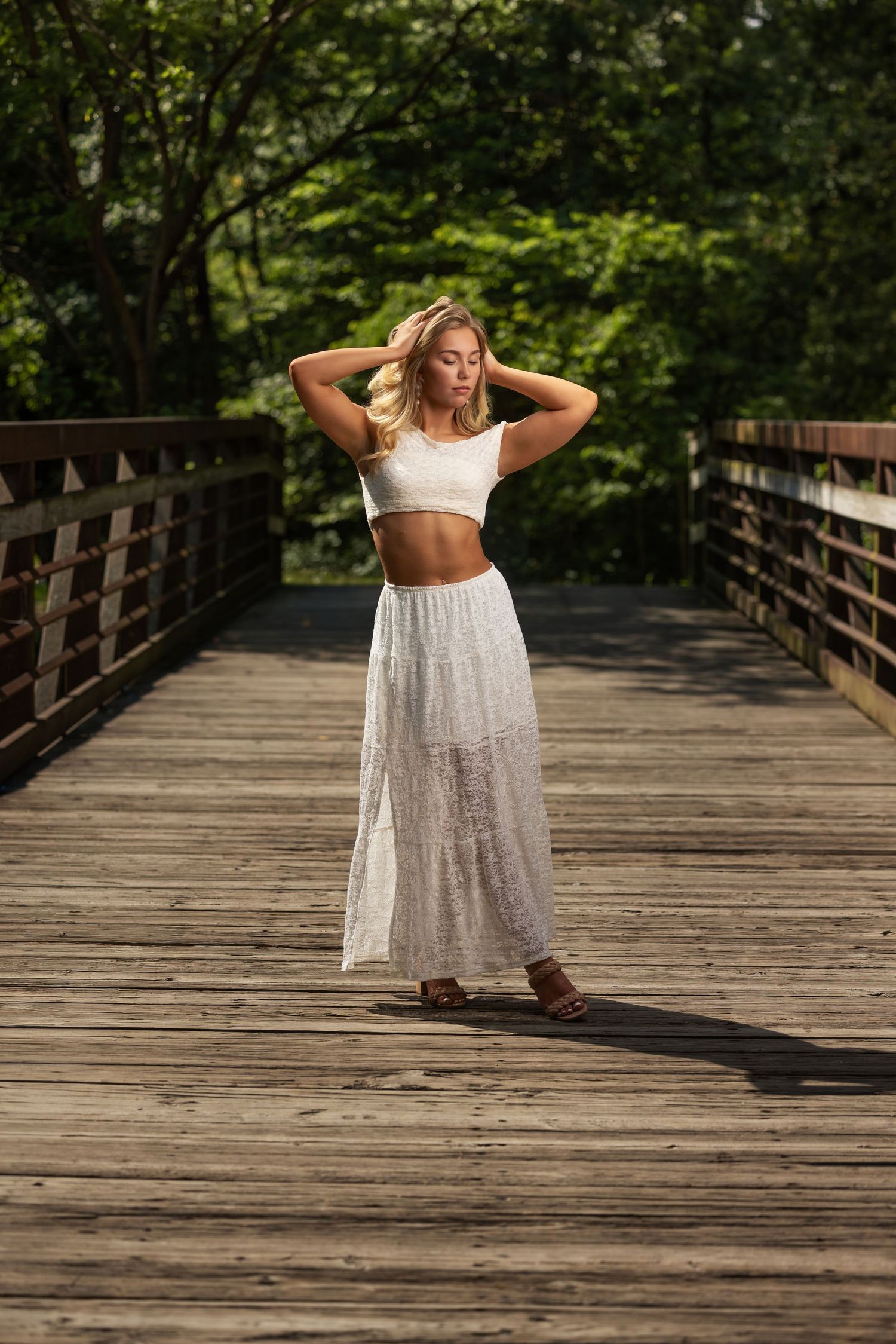 Senior portrait of a blonde girl in a flowing white dress standing on a wooden bridge
