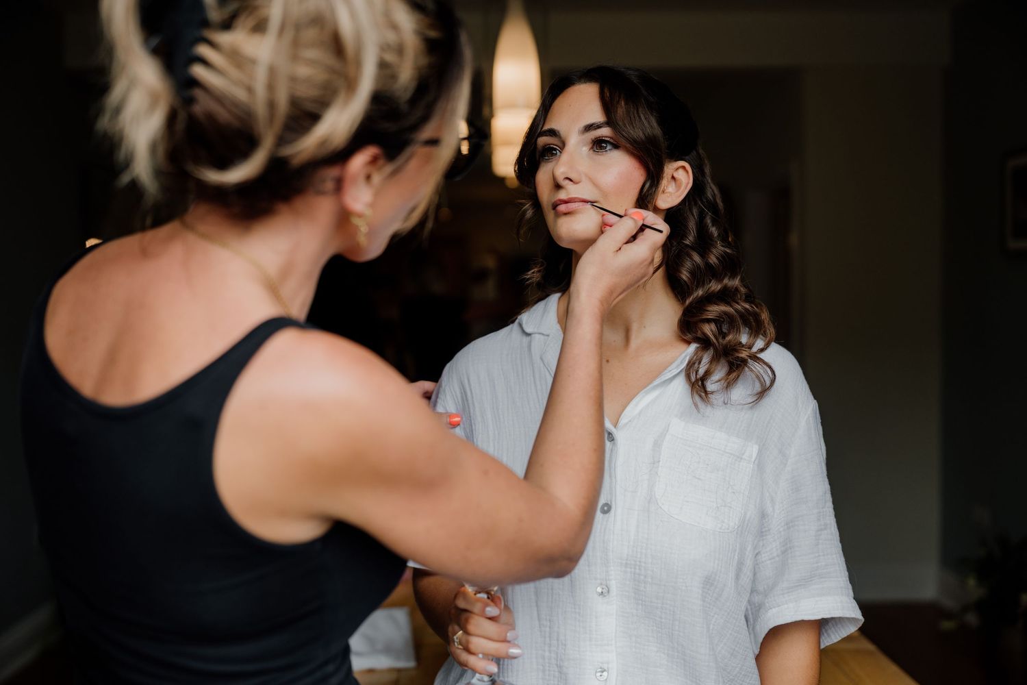 A makeup artist applies lip liner to a brunette bride in a white shirt during wedding preparations.