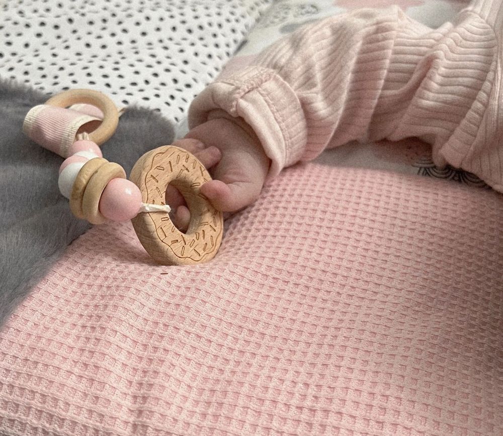 Baby hand holding a wooden donut teether rattle on a pink waffle blanket.