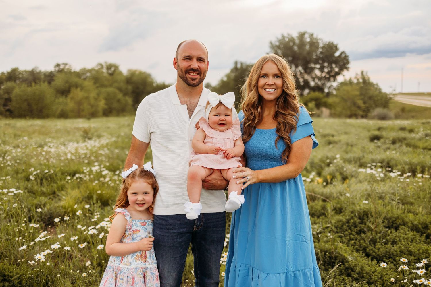A happy family of four poses in a wildflower field, with parents holding two young daughters in summer outfits.