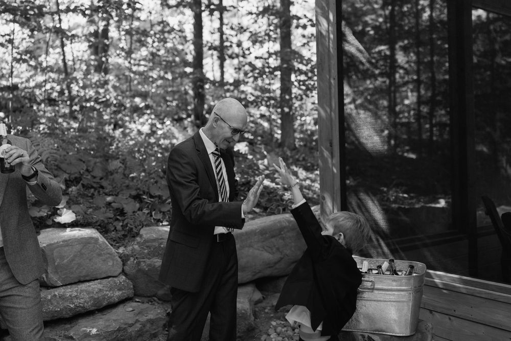 A man in a suit shakes hands with another person outdoors near stone walls and trees in a black and white photo.