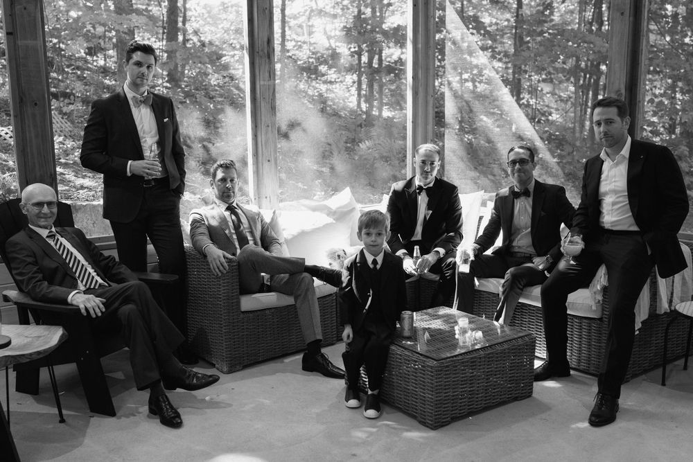 Black and white photo of a group of men in formal suits gathered in a modern glass-walled room surrounded by trees.