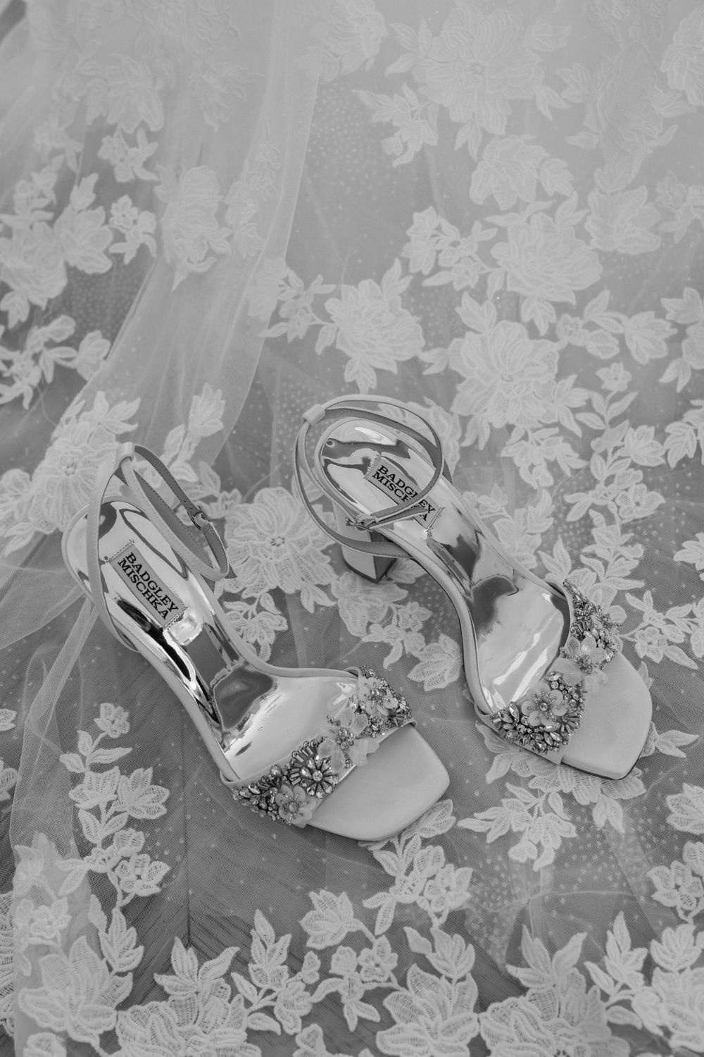 Black and white photo of Badgley Mischka bridal heels with crystal embellishments resting on floral lace wedding veil.