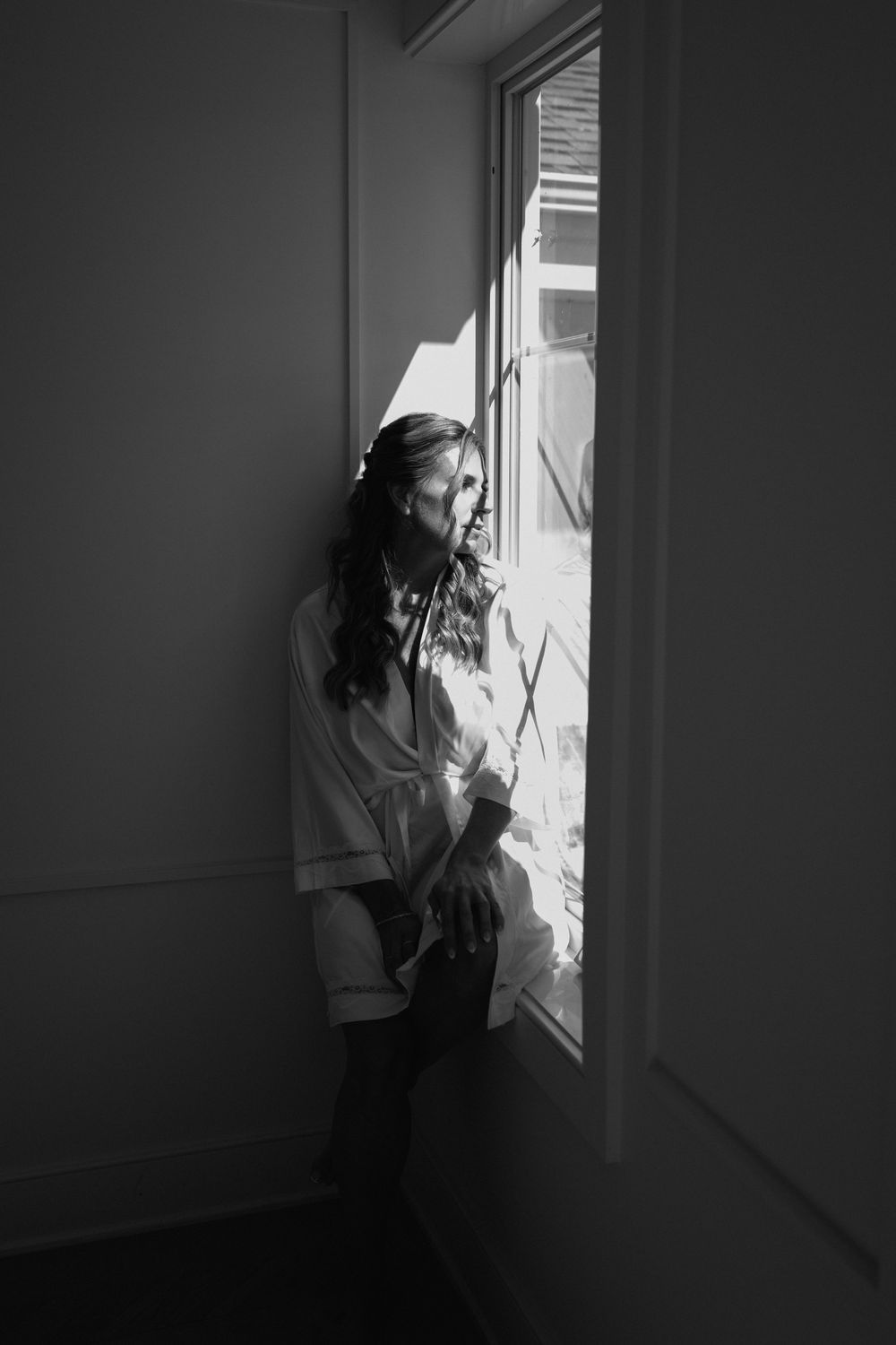 Black and white photo of a woman in a white robe standing by a sunlit window, gazing outside thoughtfully.
