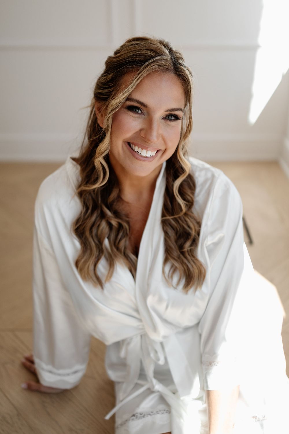 Smiling bride in white satin robe with curly highlighted hair, sitting on hardwood floor in bright natural light.