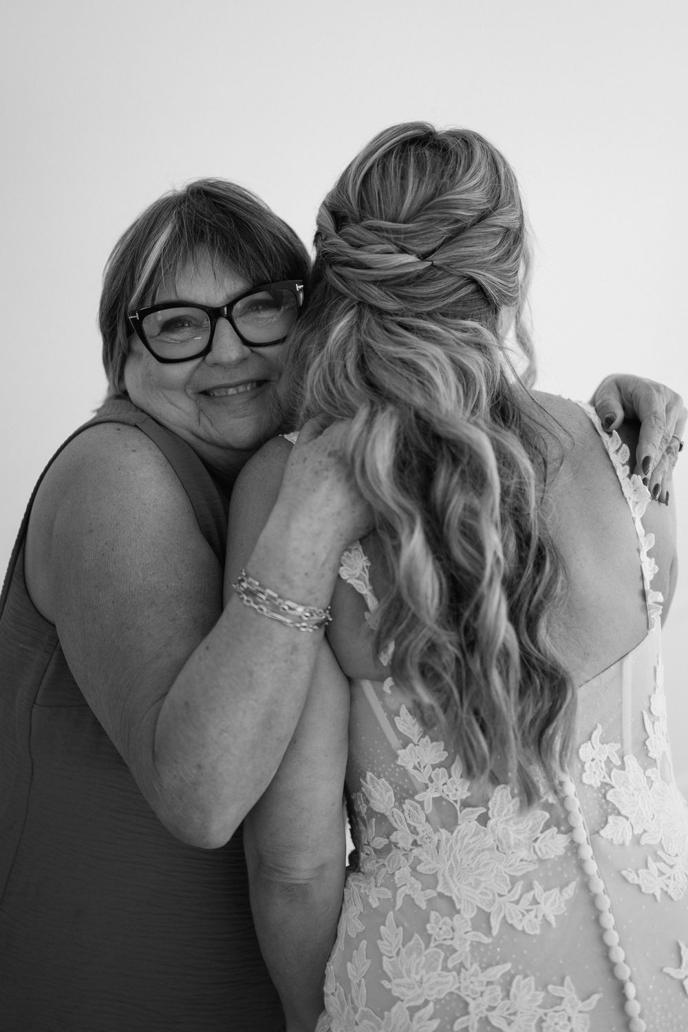 Black and white photo of a smiling mother hugging her daughter in a lace wedding dress from behind on her wedding day.
